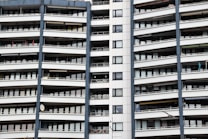 A modern apartment building with multiple floors, each featuring balconies with railings. The building has a repetitive architectural style with a mix of glass windows and white panels separated by vertical gray columns.