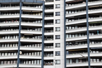 A modern apartment building with multiple floors, each featuring balconies with railings. The building has a repetitive architectural style with a mix of glass windows and white panels separated by vertical gray columns.