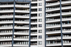 A modern apartment building with multiple floors, each featuring balconies with railings. The building has a repetitive architectural style with a mix of glass windows and white panels separated by vertical gray columns.