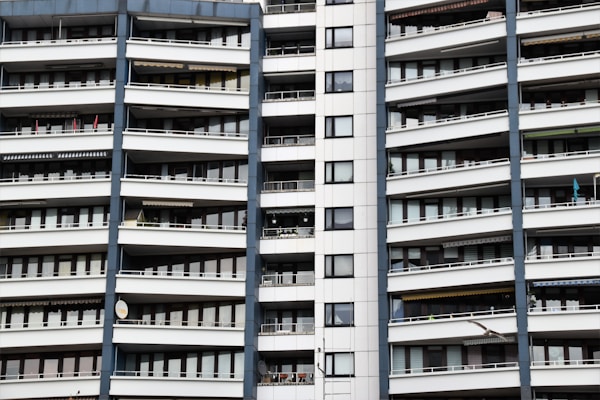 A modern apartment building with multiple floors, each featuring balconies with railings. The building has a repetitive architectural style with a mix of glass windows and white panels separated by vertical gray columns.