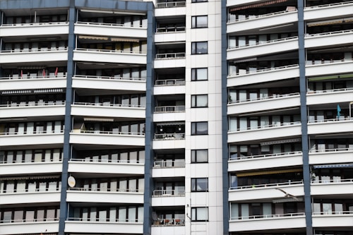 A modern apartment building with multiple floors, each featuring balconies with railings. The building has a repetitive architectural style with a mix of glass windows and white panels separated by vertical gray columns.