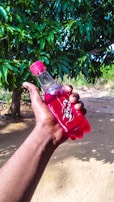 A hand is holding a red bottle labeled 'Sting' outdoors, with lush green foliage in the background. The scene is bright, suggesting a sunny day.