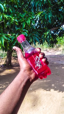 A hand is holding a red bottle labeled 'Sting' outdoors, with lush green foliage in the background. The scene is bright, suggesting a sunny day.