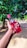 A hand is holding a red bottle labeled 'Sting' outdoors, with lush green foliage in the background. The scene is bright, suggesting a sunny day.