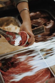 A hand scooping a portion of red-colored gelato or ice cream from a metal tray into a cup. The setting appears to be an ice cream or gelato shop, with various flavors displayed in containers. There is a small chalkboard sign in the background.