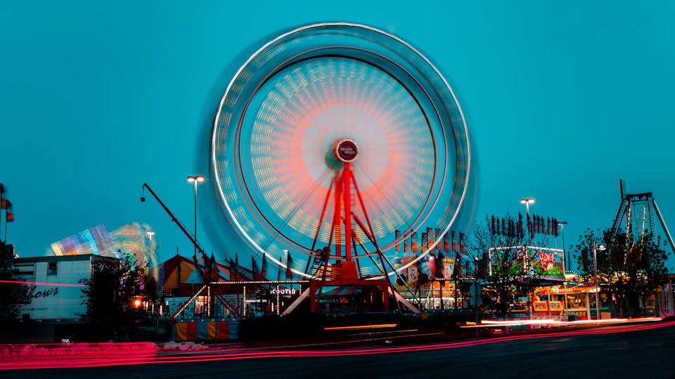 The image depicts a large, brightly lit Ferris wheel at an amusement park. The Ferris wheel is adorned with colorful lights and stands out against the dark blue sky. In the background, there are other attractions such as a carousel and a roller coaster, adding to the festive atmosphere of the scene.
