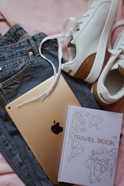 A pair of white sneakers with brown accents, a travel book with a geometric world map design, an Apple iPad, and a pair of blue jeans are laid out on a pink fabric background.