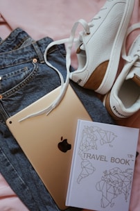 A pair of white sneakers with brown accents, a travel book with a geometric world map design, an Apple iPad, and a pair of blue jeans are laid out on a pink fabric background.