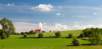 Vast green fields under a bright blue sky with scattered clouds. A red-roofed church building with a steeple sits on a gentle hill in the distance, surrounded by small trees and bushes. Bright sunlight illuminates the landscape, casting soft shadows on the grass.