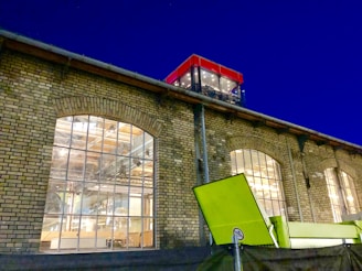 A brick industrial building with large arched windows illuminated from the inside, showcasing the interior structure and machinery. A bright green structure is visible in the foreground, while a modern glass extension with a red frame is situated on the rooftop.