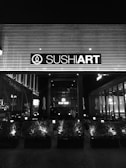 The image depicts the exterior of a sushi restaurant at night, with a prominent sign displaying the establishment's name, illuminated against a backdrop of horizontal lines. The front area features a series of planters with small bushes, and inside the restaurant, tables and chairs are visible through large windows.