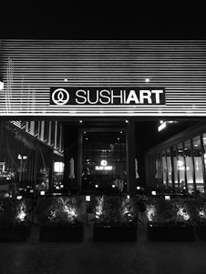 The image depicts the exterior of a sushi restaurant at night, with a prominent sign displaying the establishment's name, illuminated against a backdrop of horizontal lines. The front area features a series of planters with small bushes, and inside the restaurant, tables and chairs are visible through large windows.