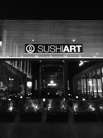 The image depicts the exterior of a sushi restaurant at night, with a prominent sign displaying the establishment's name, illuminated against a backdrop of horizontal lines. The front area features a series of planters with small bushes, and inside the restaurant, tables and chairs are visible through large windows.