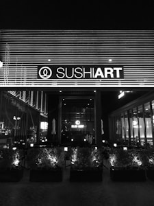 The image depicts the exterior of a sushi restaurant at night, with a prominent sign displaying the establishment's name, illuminated against a backdrop of horizontal lines. The front area features a series of planters with small bushes, and inside the restaurant, tables and chairs are visible through large windows.