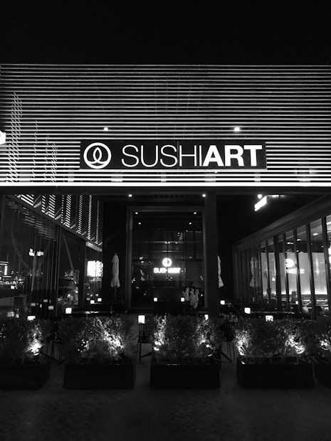 The image depicts the exterior of a sushi restaurant at night, with a prominent sign displaying the establishment's name, illuminated against a backdrop of horizontal lines. The front area features a series of planters with small bushes, and inside the restaurant, tables and chairs are visible through large windows.
