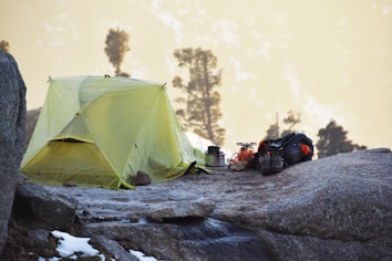A yellow tent is pitched on a rocky surface with a few patches of snow. Nearby, there is camping gear including a backpack, a pair of boots, a portable stove, and cookware. In the background, the image features trees and a mountainous landscape bathed in a soft, warm light.