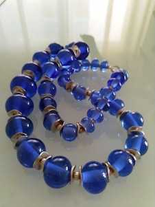 A string of large, glossy blue beads arranged in a looping pattern on a reflective surface. The beads are separated by small metallic spacers and have a semi-transparent quality, casting subtle reflections on the surface beneath.