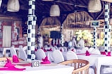 The interior of a restaurant decorated with white tablecloths and red napkins set on tables. The restaurant features distinctive black and white checkered columns, wicker light fixtures, and a wooden roof. Blurred figures in white clothing are visible in the background, indicating a staff presence.
