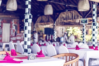 The interior of a restaurant decorated with white tablecloths and red napkins set on tables. The restaurant features distinctive black and white checkered columns, wicker light fixtures, and a wooden roof. Blurred figures in white clothing are visible in the background, indicating a staff presence.