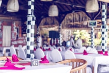 The interior of a restaurant decorated with white tablecloths and red napkins set on tables. The restaurant features distinctive black and white checkered columns, wicker light fixtures, and a wooden roof. Blurred figures in white clothing are visible in the background, indicating a staff presence.