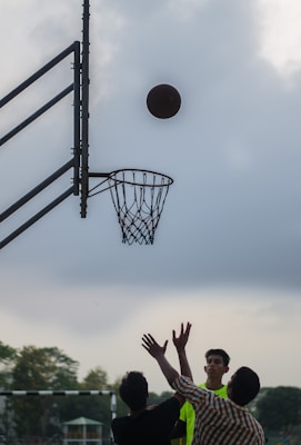 A basketball is about to go through a hoop during an outdoor game. Three people are gathered below the hoop, one of them wearing a bright yellow shirt. The sky is overcast, adding a dramatic feel to the scene.
