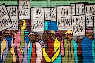 A colorful mural depicting a group of people holding signs that read 'I AM A MAN.' The artwork is painted on a brick wall, with people wearing various vibrant colored clothing, including hues of blue, pink, yellow, and green.