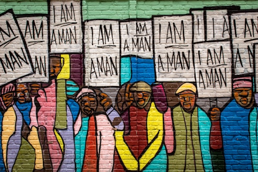 A colorful mural depicting a group of people holding signs that read 'I AM A MAN.' The artwork is painted on a brick wall, with people wearing various vibrant colored clothing, including hues of blue, pink, yellow, and green.