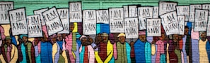 A mural depicting a group of people holding signs that read 'I Am A Man'. The figures are diverse, with a variety of clothing styles and colors. The background is composed of brick, and the mural uses vibrant, bold colors, emphasizing unity and strength.