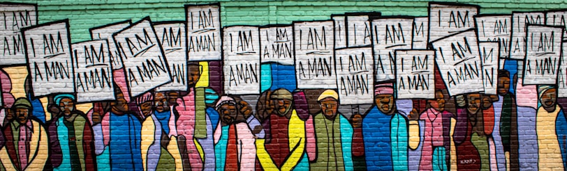 A mural depicting a group of people holding signs that read 'I Am A Man'. The figures are diverse, with a variety of clothing styles and colors. The background is composed of brick, and the mural uses vibrant, bold colors, emphasizing unity and strength.
