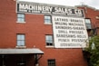 A brick building features multiple windows and a large painted sign advertising a machinery sales company. The sign lists various types of metalworking machinery, including lathes, brakes, milling machines, and more. An external metal staircase runs along the side of the building with some greenery visible at the bottom right.