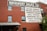 A brick building features multiple windows and a large painted sign advertising a machinery sales company. The sign lists various types of metalworking machinery, including lathes, brakes, milling machines, and more. An external metal staircase runs along the side of the building with some greenery visible at the bottom right.