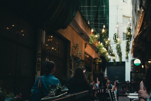 A cozy, narrow alleyway with an intimate dining area. Overhead, strings of lights and hanging plants create a warm, inviting atmosphere. People are seated at tables, engaged in conversation. The lighting is dim, with soft, moody tones.