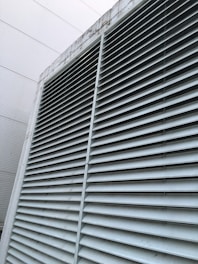 A large industrial vent is featured prominently, with horizontal metal slats covering its entirety. The structure appears to be part of a building exterior, with visible weathering and discoloration on the surfaces.