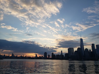 The image displays a city skyline at sunset with towering skyscrapers silhouetted against a sky filled with scattered clouds. The water in the foreground reflects the dark shapes of the buildings and the colorful sky.