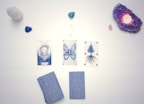 Three tarot cards are arranged in a row on a white surface, depicting a lion, a butterfly, and a tree with roots. Surrounding the cards are several crystals, including an amethyst cluster with a lit candle and various other gemstones.