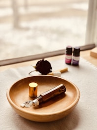 A wooden bowl contains an amber-colored glass bottle with a metallic roller ball and a gold cap. Small white stones are placed beside the bottle. In the background, two small bottles with purple and pink labels are visible near a dark dried flower, all placed on a light-colored fabric surface near a window.