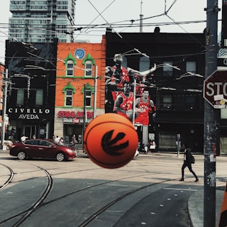A busy urban street scene features a large basketball in the foreground with a sports team logo. Behind the ball, a wall mural depicts several basketball players in red jerseys. The street is lined with shops, including one with a distinct orange facade and green trim. Overhead, multiple trolley cables crisscross the sky. A red sedan is driving on the street, and pedestrians are walking in different directions.