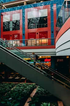 A person with a backpack is ascending an escalator inside a modern shopping mall. The background features bright red and blue geometric patterns on a storefront, identified as a pharmacy and wellness store. Lush greenery decorates the area below the escalator, providing a contrast to the sleek, industrial architecture.