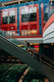 A person with a backpack is ascending an escalator inside a modern shopping mall. The background features bright red and blue geometric patterns on a storefront, identified as a pharmacy and wellness store. Lush greenery decorates the area below the escalator, providing a contrast to the sleek, industrial architecture.