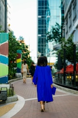 A person wearing a bright blue graduation gown walking along a city sidewalk, surrounded by modern buildings and greenery. The perspective captures the urban landscape with reflections on a glass building and colorful panels along the path.