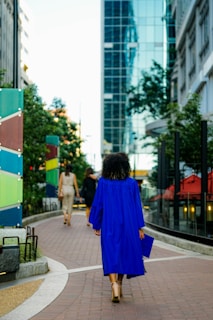 A person wearing a bright blue graduation gown walking along a city sidewalk, surrounded by modern buildings and greenery. The perspective captures the urban landscape with reflections on a glass building and colorful panels along the path.