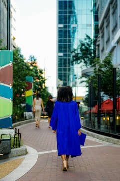A person wearing a bright blue graduation gown walking along a city sidewalk, surrounded by modern buildings and greenery. The perspective captures the urban landscape with reflections on a glass building and colorful panels along the path.