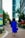 A person wearing a bright blue graduation gown walking along a city sidewalk, surrounded by modern buildings and greenery. The perspective captures the urban landscape with reflections on a glass building and colorful panels along the path.