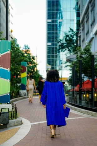 A person wearing a bright blue graduation gown walking along a city sidewalk, surrounded by modern buildings and greenery. The perspective captures the urban landscape with reflections on a glass building and colorful panels along the path.