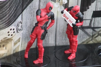 Two red and black figurines resembling comic book characters are displayed. One figure holds a sign with humorous text while the other appears to be posing thoughtfully. The background includes film reels and industrial decor.