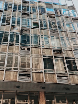 A close-up view of a building facade consisting of numerous glass windows and metal frames. Some windows have air conditioning units installed. The glass reflects various patterns and distortions of surrounding structures.
