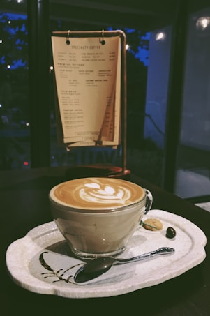 A latte with artfully crafted milk foam is served in a clear glass cup on a patterned ceramic tray. Alongside sits a small cookie, a coffee bean, and a spoon. A menu on a wooden stand in the background displays various coffee options, set against a dimly lit interior.