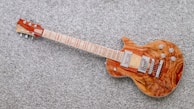 A wooden electric guitar with a natural finish rests on a textured gray carpet. It features a glossy body with intricate wood grain patterns and metallic hardware components such as pickups and control knobs. The guitar neck contains distinct red fret markings.