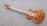 A wooden electric guitar with a natural finish rests on a textured gray carpet. It features a glossy body with intricate wood grain patterns and metallic hardware components such as pickups and control knobs. The guitar neck contains distinct red fret markings.