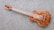 A wooden electric guitar with a natural finish rests on a textured gray carpet. It features a glossy body with intricate wood grain patterns and metallic hardware components such as pickups and control knobs. The guitar neck contains distinct red fret markings.